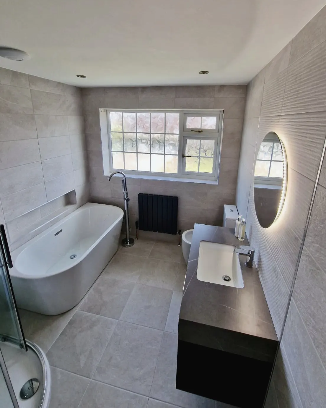 Bathroom in warm stone-effect tiles with a freestanding bath beneath the window and a wall-hung basin